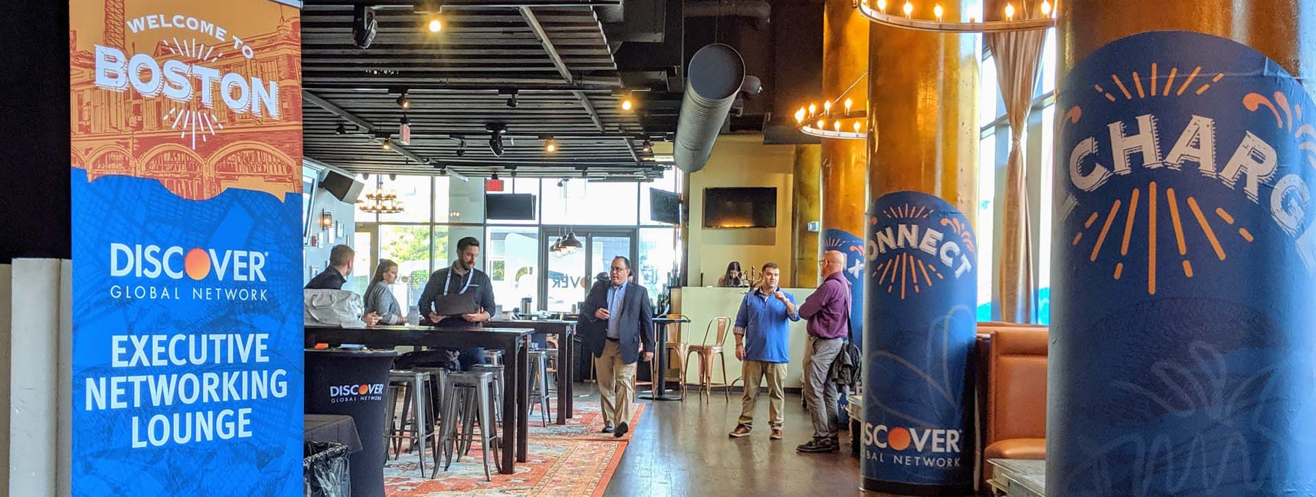 A pop-up banner welcomes guests to the Discover Networking Lounge, an open space with charging tables, orange rugs and vinyl wrapped columns