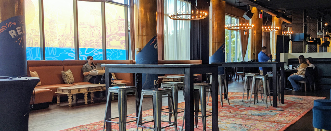 An image of the open lounge area with wooden charging tables, metal barstools, blue velvet ottomans sit atop a vibrant orange rug