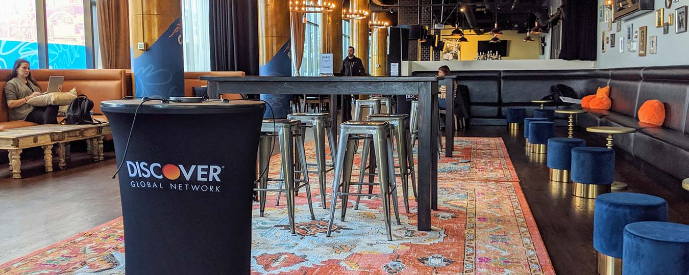 close-up image of a black highboy table branded with Discover logo and cables atop for charging devices, in the background are wooden bar tables atop a vibrant orange rug with metal barstools, blue velvet ottomans and gold side tables line black leather seating along the wall