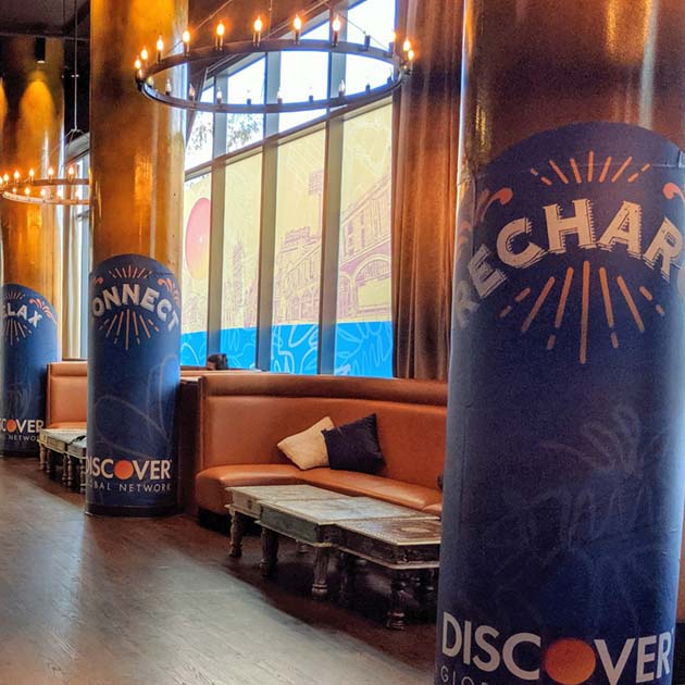 close-up of three architectural columns wrapped in blue adhesive vinyl, each labeled "Recharge", "Connect", and "Relax". In between each column are tan leather booths along windows covered in a transparent vinyl with a Boston skyline graphic applied.