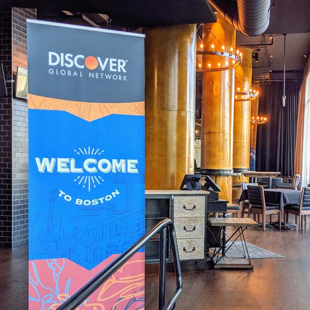 A close-up image of a vinyl pop-up banner, the top is black with the Discover logo, a blue middle section reads "Welcome to Boston" in a vintage letterpress style typeface, the lower section is terracotta colored with yellow and blue organic, abstract lines