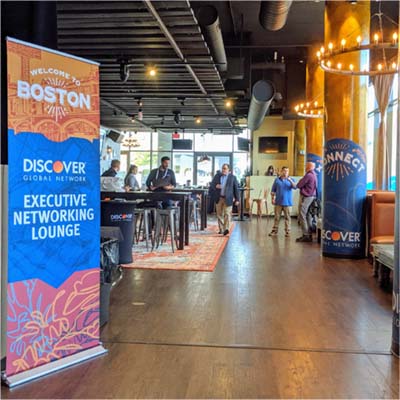 interior of a Boston bar with an industrial feel is branded orange and blue for Discover Global Network, including a pop-up banner which says "Welcome to Boston", executives gather around device charging counters and columns wrapped in blue adhesive vinyl