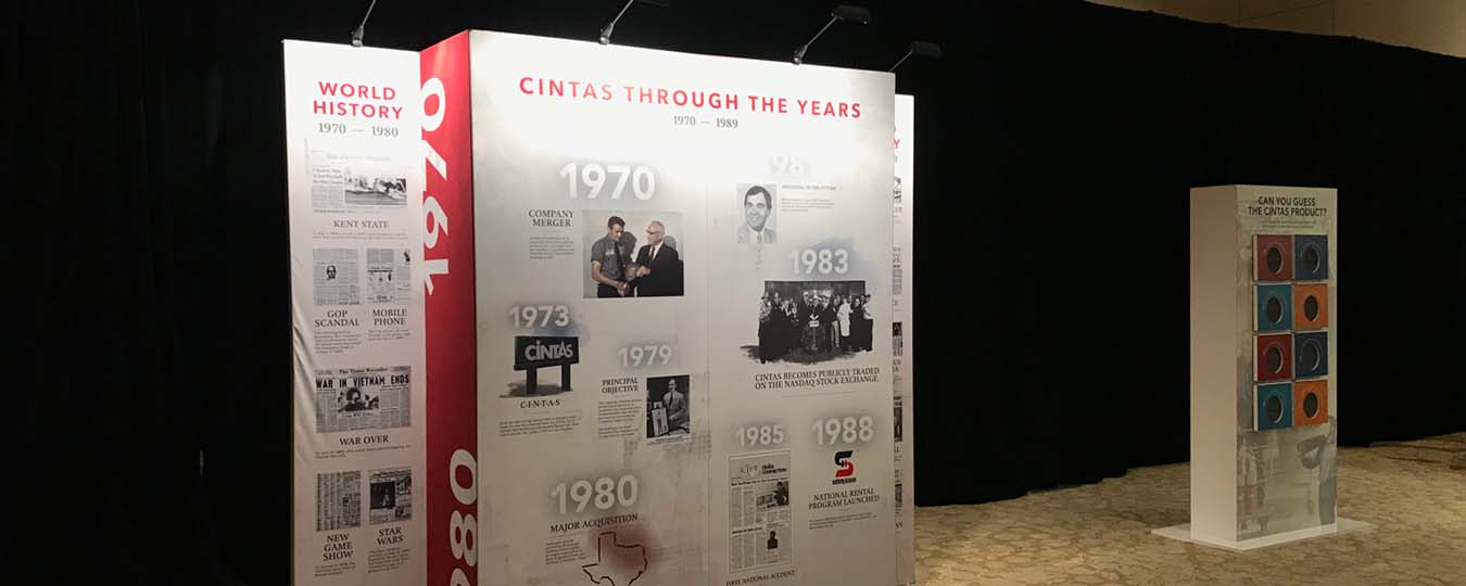 large timeline display, roughly 6 feet tall and 10 feet wide, on display in a ballroom with carpeted floors, next to another structure labeled "Can you guess the Cintas Product?" with doors with holes to reach in and feel textured products