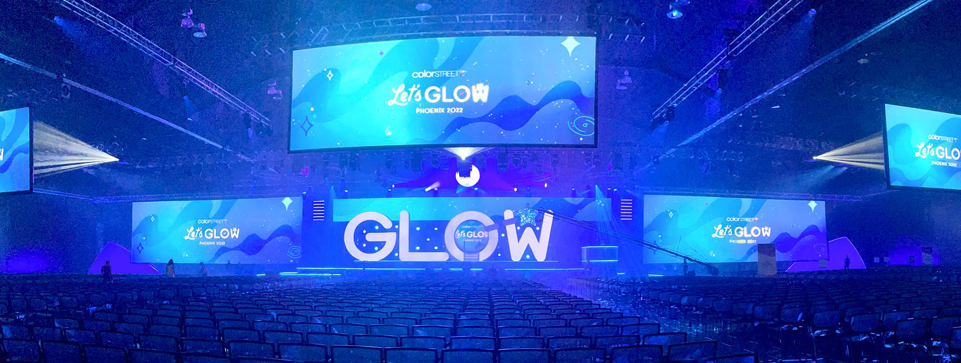 A view of a stage washed in blue lighting with oversized "GLOW" letters and several large projection screens