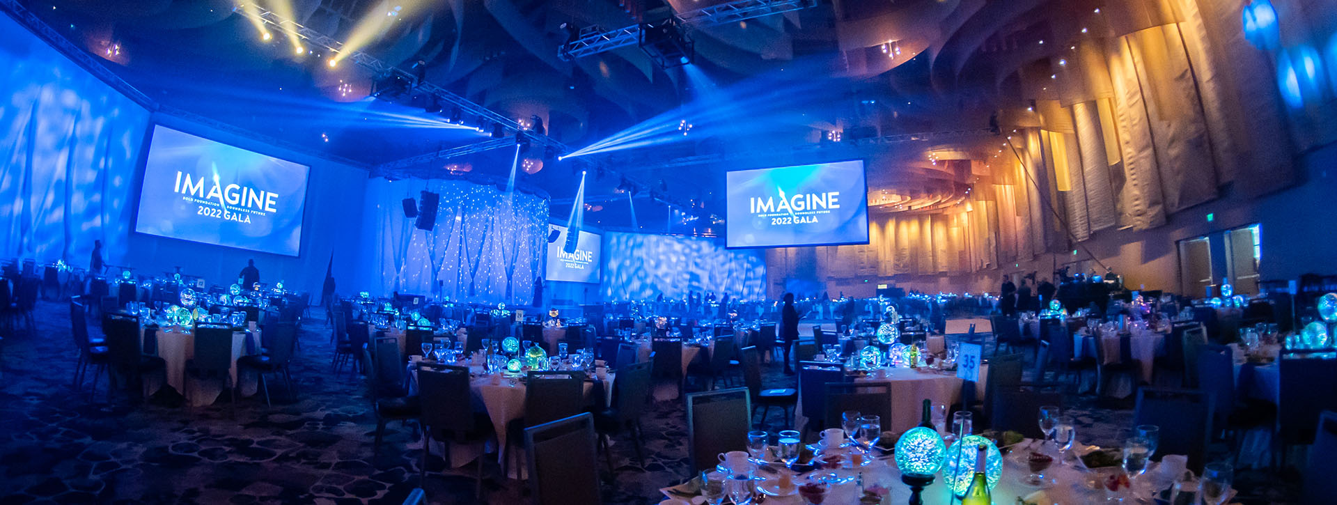 A ballroom is painted with blue and yellow light, table rounds are placed throughout the space with glowing orb centerpieces. Several projection screens show a logo reading "Imagine" and drapery lines the stage.